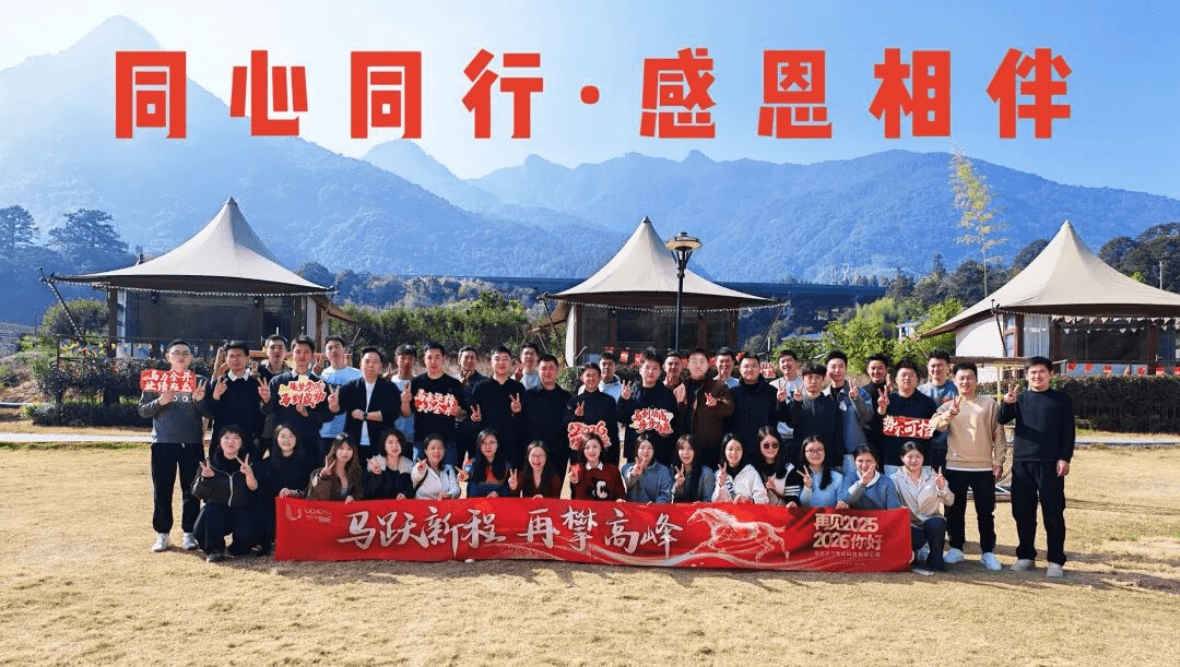 公司年会团队大合影,背景为山景和帐篷,红色横幅写着同心同行感恩相伴
