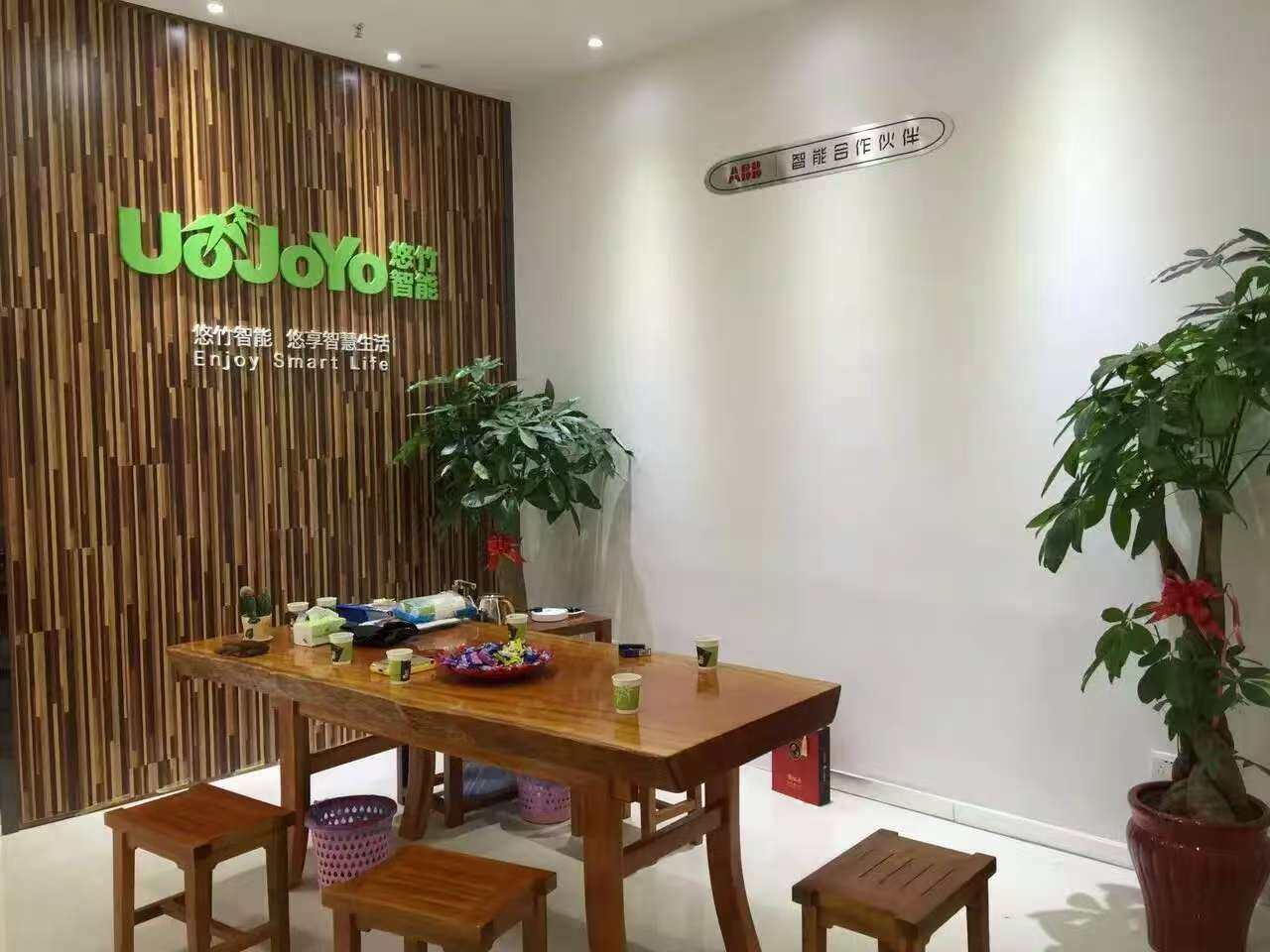 悠竹智能展厅接待区,木质条纹背景墙,绿色 UoJoYo 悠竹智能 Logo,悠竹智能您享智慧生活 Enjoy Smart Life 标语,ABB 智能合作伙伴标识,实木茶桌和绿植