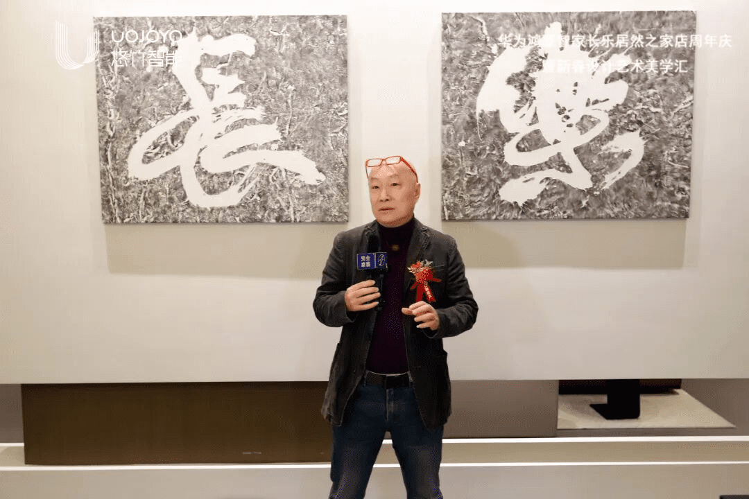 国际职业艺术家董兴接受专访,穿着深色西装和紫色毛衣,佩戴胸花,手持麦克风,背景是灰色书法作品