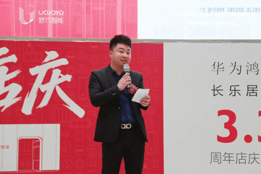 长乐店总经理穿着黑色西装,佩戴胸花,手持麦克风和讲稿在红色背景墙前致辞