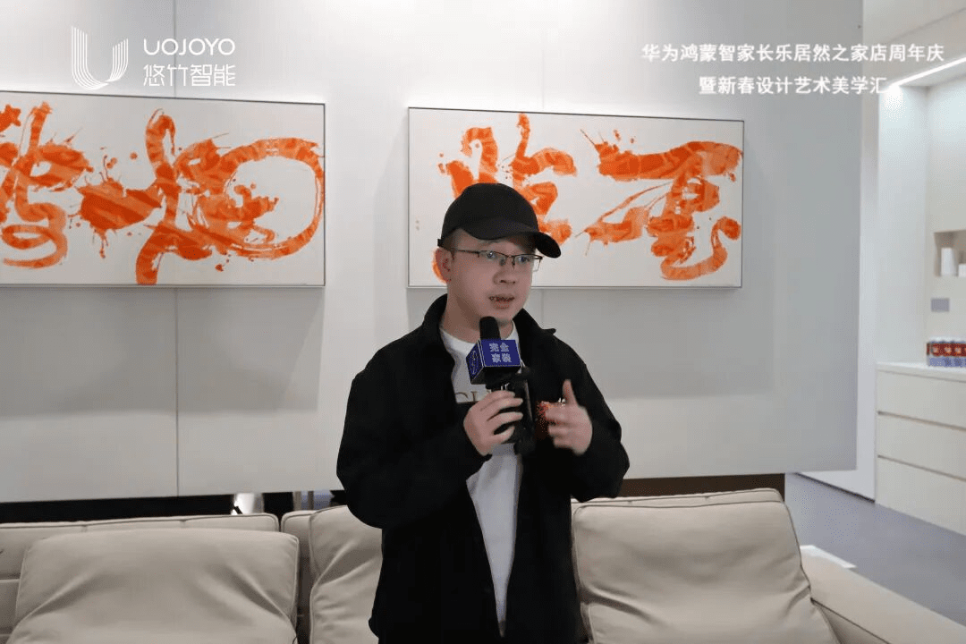 杰艾设计郑杰接受专访,穿着黑色外套戴黑色帽子,手持麦克风,背景是橙色书法作品
