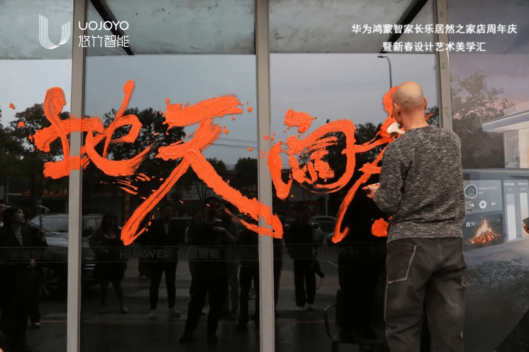 周雄波老师在玻璃墙上用橙色颜料题字广阔天地,光头穿着灰色毛衣,背对镜头创作